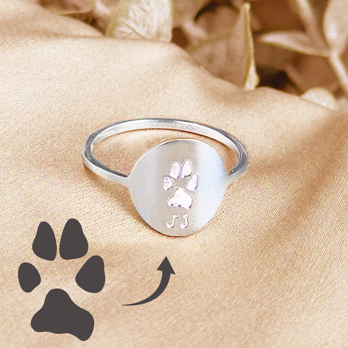 Sterling silver custom paw print ring with a brushed oval signet face engraved from a real pet paw print, shown on a soft champagne satin background with an illustrated paw icon and arrow demonstrating how your dog or cat’s paw is transformed into jewelry. Dainty minimalist pet memorial ring and dog mom gift, personalized keepsake to remember a beloved furry friend, perfect for everyday wear, birthdays, Christmas or sympathy gifts.