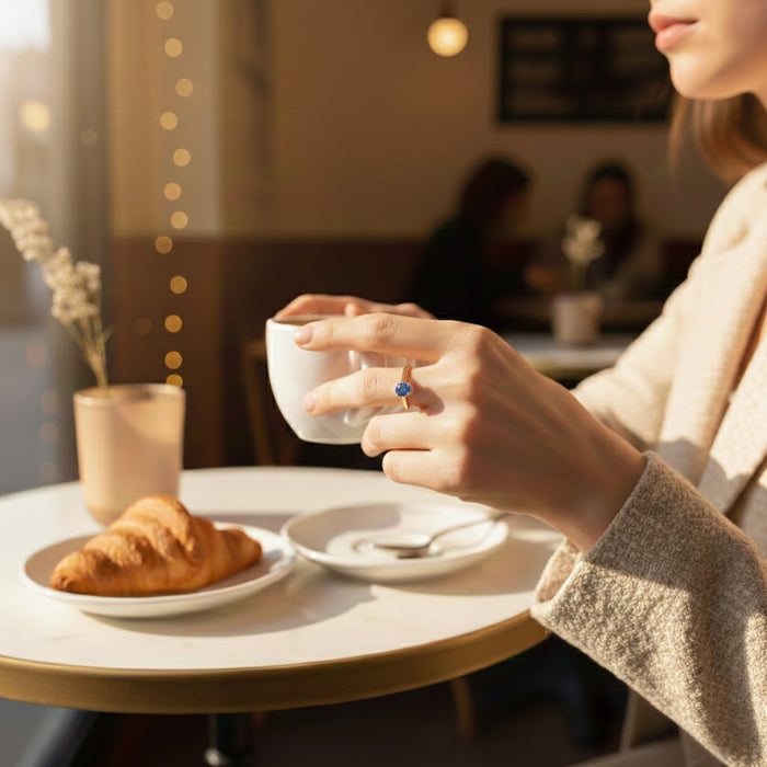 Dainty gold stackable birthstone ring with vibrant blue gemstone worn on a woman’s hand as she holds a coffee cup in a cozy café, soft morning light and bokeh lights in background, croissant and minimal table setting, thoughtful everyday mothers ring gift.