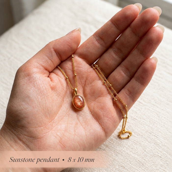 Hand holding a sunstone pendant necklace, showing the 8x10mm oval orange gemstone pendant and delicate gold chain for scale.