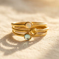 Tiny cabochon birthstone stacking rings in gold with opal, citrine, and aqua stones, shown on soft fabric as a dainty personalized ring gift.