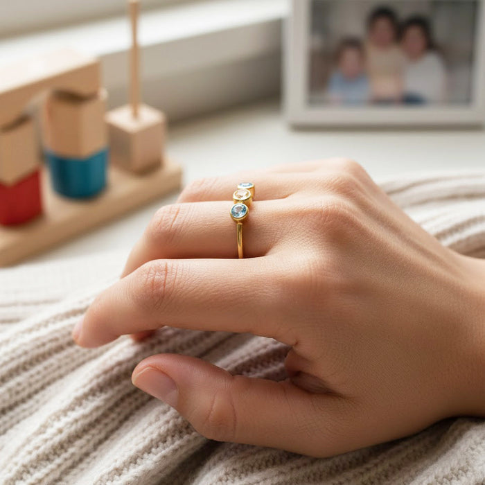 Dainty mom birthstone ring with three bezel-set gemstones in a slim gold band, worn on a woman’s hand resting on a cozy knit blanket, symbolizing three children or family members; meaningful personalized birthstone jewelry gift for mom for Mother’s Day, birthday, or everyday wear.