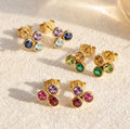 Custom triple birthstone stud earrings with three 3mm gemstones in gold settings on a soft neutral fabric background.
